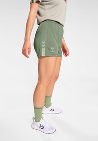 Pantalones cortos deportivos verdes con cintura ajustable, con detalles claros y un logo. Combinados con zapatillas blancas y calcetines verdes a juego.