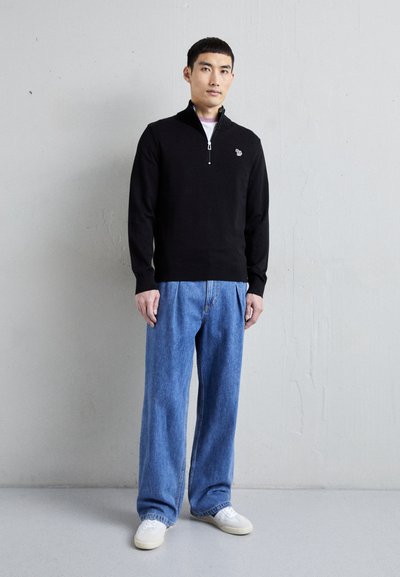 Fekete, cipzáros pulóver kötött anyagból, világoskék, bő nadrággal párosítva. A fehér sportcipők egészítik ki az öltözéket. Egyszerű hímzett logó részlet látható.