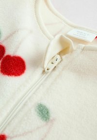 Valkoinen fleece-vaate, jossa on punaisia ja vihreitä pyöreitä kuvioita, etuvetoketjukiinnityksellä ja pehmeällä pinnalla. Tunnus näkyvissä sisällä.