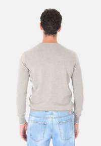 Uomo con capelli corti scuri che indossa un maglione grigio chiaro e jeans azzurri, in piedi di spalle contro uno sfondo bianco.