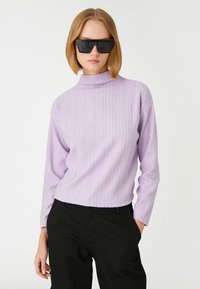 Fioletowy dzianinowy golf z teksturowanym wzorem w zygzaki, zestawiony z czarnymi spodniami. Modelka nosi odważne czarne okulary przeciwsłoneczne.