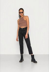 Top castanho, jeans pretos de cintura alta e botas pretas com atacadores. A pessoa está de pé à frente de um fundo neutro, com óculos de sol elegantes.