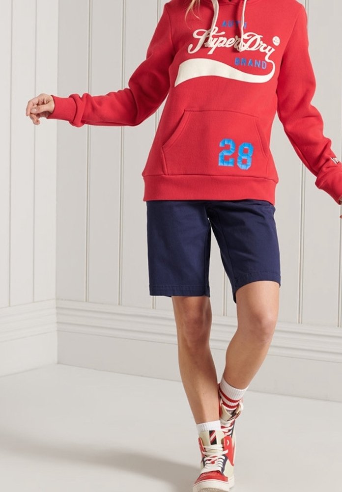 Personne portant un sweat à capuche rouge avec le texte "SuperDry", un short bleu marine, des chaussettes blanches et des baskets montantes rouges, debout à l'intérieur.
