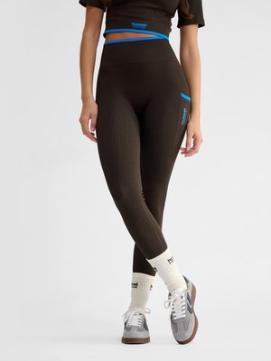 Vrouw draagt zwarte geribde legging en crop top met blauwe bies, witte sokken en grijze sneakers, staand met gekruiste benen.