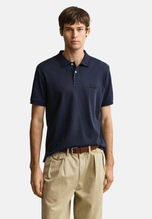 Junger Mann, der ein marineblaues, kurzärmeliges Poloshirt trägt, das in beige plissierte Hosen mit braunem Ledergürtel gesteckt ist, steht vor einem weißen Hintergrund.