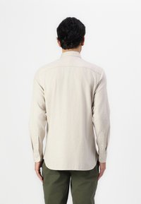 Cămașă bej cu mânecă lungă, cu guler clasic, manșete cu nasturi și burtă dreaptă, purtată cu pantaloni de culoare verde măsliniu.
