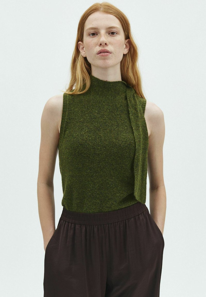 Jeune femme aux longs cheveux roux portant un haut en tricot vert sans manches et un pantalon ample foncé, debout devant un fond clair uni.