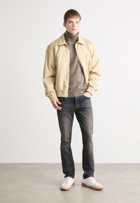Chaqueta bomber beige, suéter de punto marrón con cuello alto, vaqueros oscuros y zapatillas blancas con suelas de goma. Tejido suave con puños elásticos en la chaqueta.