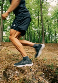 Zapatillas deportivas negras con una superficie texturada y suela blanca, combinadas con calcetines grises, exhibidas en un sendero forestal con follaje verde.