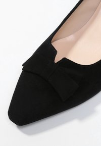 Chaussure plate en suède noire avec un bout pointu et un accent de nœud prominent à l'avant. L'intérieur est beige, mettant en valeur des contrastes de textures lisses.