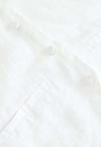 Fehér lenvászon anyag texturált felülettel, két kerek, anyaggal borított gombbal és oldalzseb részlettel.