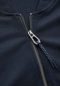 Il giubbotto zip-up navy presenta una zip in metallo con un tirante decorativo. Il tessuto ha una consistenza liscia e un design del colletto aderente.