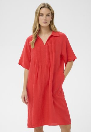 Mujer con un vestido rojo suelto, de manga corta, hasta la rodilla, con cuello y plisados verticales en la parte delantera, de pie con una mano en el bolsillo.