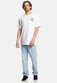 Weißes Baumwoll-T-Shirt mit kurzen Ärmeln und rundem Halsausschnitt, mit einem runden schwarzen Logo auf der linken Brust. Hellblaue Jeans und schwarze Sneakers.