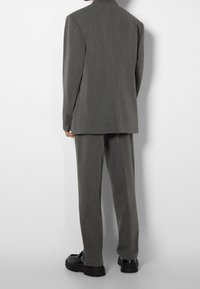 En person står med ryggen mot kameran, iklädd en grå kostym med matchande byxor och svarta kraftiga skor, mot en enkel bakgrund.