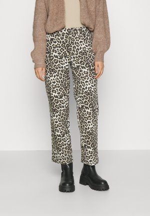 Personne portant un pantalon à motif léopard, un cardigan marron en maille et des bottines noires épaisses, debout devant un fond uni.