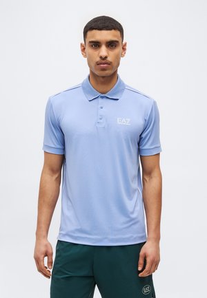 Fiatal férfi rövid hajjal, világoskék EA7 pólót és sötétzöld EA7 rövidnadrágot viselve, egyszerű fehér háttér előtt áll.