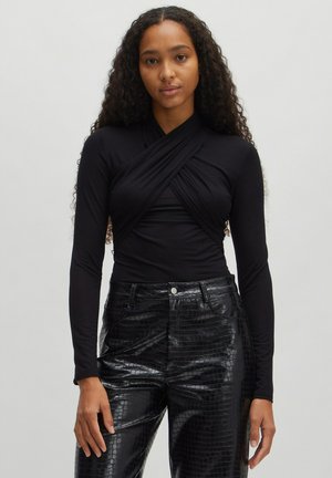 Schwarzes Langarmoberteil mit Cross-Wrap-Design, kombiniert mit hochgeschnittenen schwarzen Kunstlederhosen mit strukturiertem Krokodilmuster.