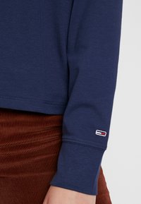 Námořnická modrá košile s dlouhým rukávem vyrobená z měkké bavlny s žebrovanými manžetami; na rukávu má malou vyšitou nášivku. Pod tímto oblečením se nacházejí kalhoty z manšestru v barvě rzi.