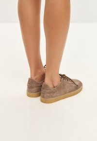 Zapatillas de ante marrón con un perfil bajo, puntera redondeada y cierre de cordones. Presentan una parte superior texturizada y una suela de goma en tono beige.