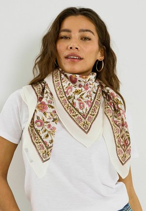 Mujer con cabello castaño ondulado, lleva grandes pendientes de aro, una camiseta blanca y una bufanda blanca con estampado floral colgada alrededor de su cuello.