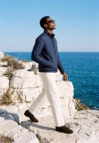 Homme portant des lunettes de soleil, une veste matelassée bleu marine, un pantalon blanc et des chaussures marron marchant sur des rochers blancs au bord de la mer sous un ciel bleu clair.