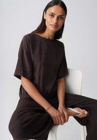 Woman with long dark hair sitting on white chair, wearing a loose dark brown top and pants, holding one foot with white flip-flop sandal.