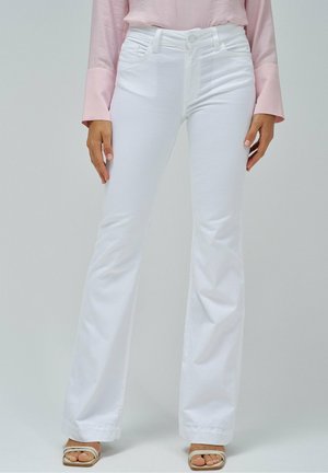 Kobieta ubrana w białe dzwoniaste dżinsy, blado różową bluzkę z długim rękawem i beżowe sandały z odkrytymi palcami, stojąca na tle jasnego, gładkiego tła.