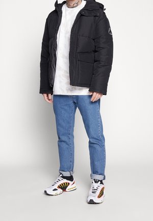 Chaqueta de plumas negra con capucha, cremallera y dos bolsillos frontales. Llevada sobre una camiseta blanca con un patrón abstracto y un pantalón vaquero azul claro. Zapatillas blancas.