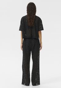 Svart spets tvådelad outfit som har en löst sittande topp med korta ärmar och vida byxor, med geometriska mönster och genomskinlighet.