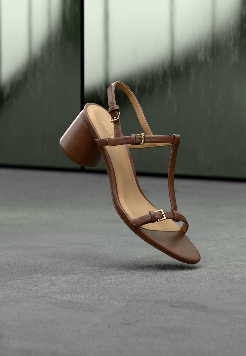 Καφέ δερμάτινο γυναικείο σανδάλι με χοντρό τακούνι και δύο λουριά με χρυσές αγκράφες, να αιωρείται πάνω από μια γκρίζα επιφάνεια με αντανάκλαση στο φόντο.