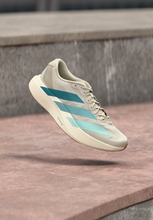 Chaussure de course beige clair avec trois bandes bleues, semelle blanche et lacets, montrée en l'air au-dessus d'une surface en béton.