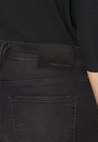 Jeans de mezclilla negra con una textura suave. Presenta un parche rectangular de cuero negro en la cintura con la inscripción "TRUE RELIGION" en relieve.