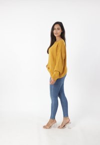 Mosterdgele gebreide trui met verlaagde schouders, gecombineerd met blauwe skinny jeans en transparante blokhak-sandalen, tegen een witte achtergrond.