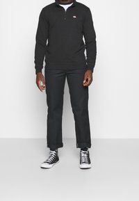 Pullover nero con colletto a mezzo zip e logo ricamato, abbinato a pantaloni neri e sneaker alte nere. Texture del tessuto liscia e casual.