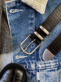 Melns teksturēts ādas josta ar sudraba divdabīgu sprādzi, kopā ar gaiši ziliem džinsiem ar bārkstīm un pelēku adītu džemperi.