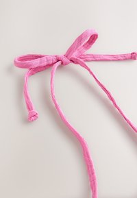 Rozā auduma lenta ar fakturētu virsmu, kurai ir mezglots loks un divi gari, plati gali. Krāsa ir spilgta un vienmērīga.