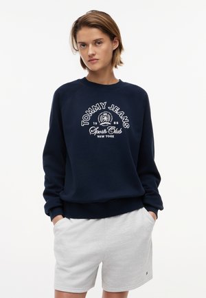 Νεαρό άτομο που φοράει φούτερ Tommy Jeans Sports Club σε ναυτικό μπλε και ανοιχτό γκρι σορτς, στέκεται με τα χέρια στις τσέπες, μπροστά σε λευκό φόντο.