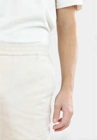 Ljusbeige bomullbyxor med elastisk midja, matchade med en vit T-shirt med kort ärm. Materialet verkar mjukt med en jämn textur.