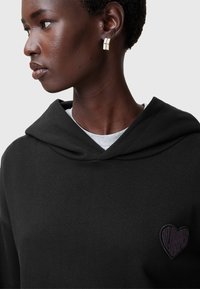 Felpa nera con una texture morbida, caratterizzata da una toppa a forma di cuore viola in contrasto sul lato sinistro del petto e un design del cappuccio rilassato e drappeggiato.