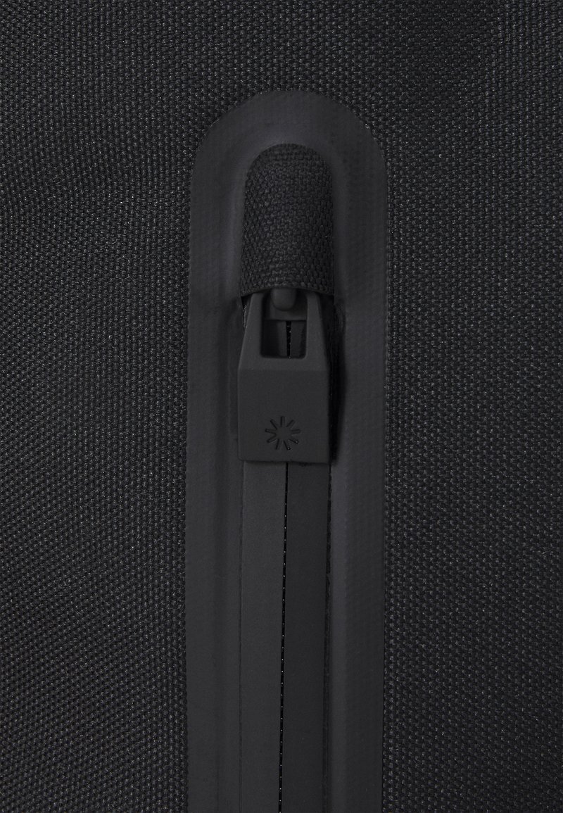 Sac en tissu noir avec une surface texturée. Présente une fermeture éclair noire avec un tirette de marque, intégrée au bord supérieur pour une fermeture sécurisée.