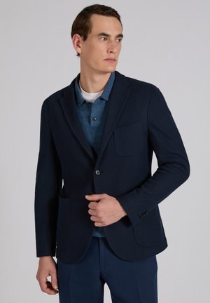 Man in een marineblazer over een blauw poloshirt en marineblauwe broek, kijkend naar de zijkant tegen een effen grijze achtergrond.
