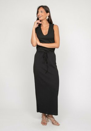 Femme souriante posant avec la main près du visage, portant une robe noire sans manches avec taille à cordon et des sandales dorées à talons hauts et à lanières.