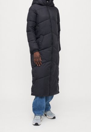 Persoon die een lange zwarte gewatteerde jas draagt met een voorrits, zijdrukknopen, twee zakken, blauwe spijkerbroek en grijze sneakers, staand op een witte achtergrond.