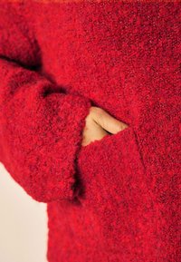 Rød struktureret sweater med løs pasform, der har en sidelomme og brede manchetter lavet af blødt, fuzzy materiale. Nærbillede.
