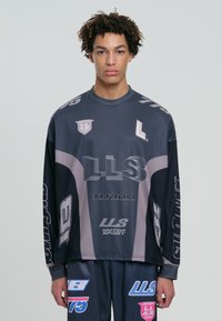 Tânăr cu păr creț, îmbrăcat cu o cămașă cu mâneci lungi în nuanțe de navy și gri, și pantaloni asortați, decorat cu logo-uri și texte de stil motorsport.