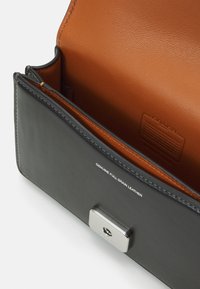 Černá taška z plné hovězí kůže s uzávěrem na sponu, oranžový kožený interiér a embosovaná značka uvnitř. Hladká textura a neat detail šití.