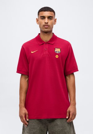 Młody mężczyzna w czerwonej koszulce polo FC Barcelona z logo Nike i Spotify, stojący na jednolitym jasnym tle.