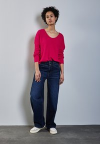 Top a maniche lunghe rosa brillante con scollatura ampia abbinato a jeans larghi blu scuro. Sneakers beige completano l'outfit.