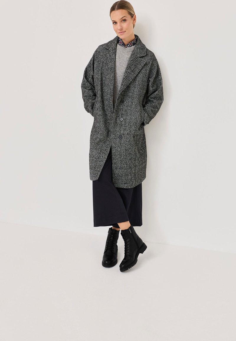 Manteau en mélange de laine gris avec un design structuré, grand revers et deux boutons, superposé sur un pull gris, associé à des bottines noires.